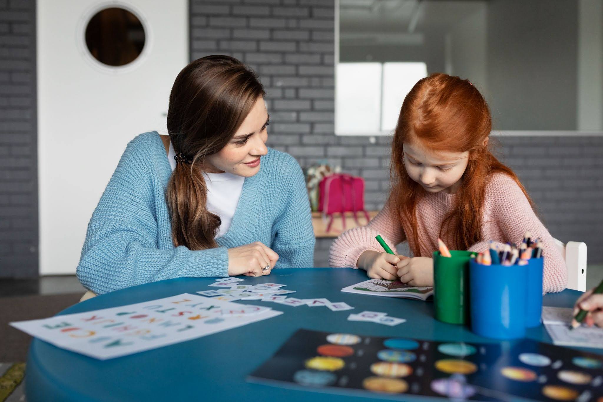 A female teacher helping a red haired girl at a table.