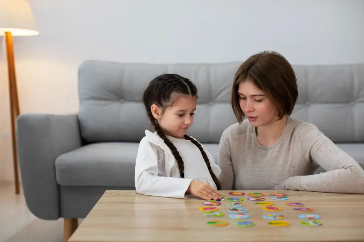 A woman and a young girl sit at a wooden table focusing on an arrangement of colorful round memory matching cards with animal drawings.