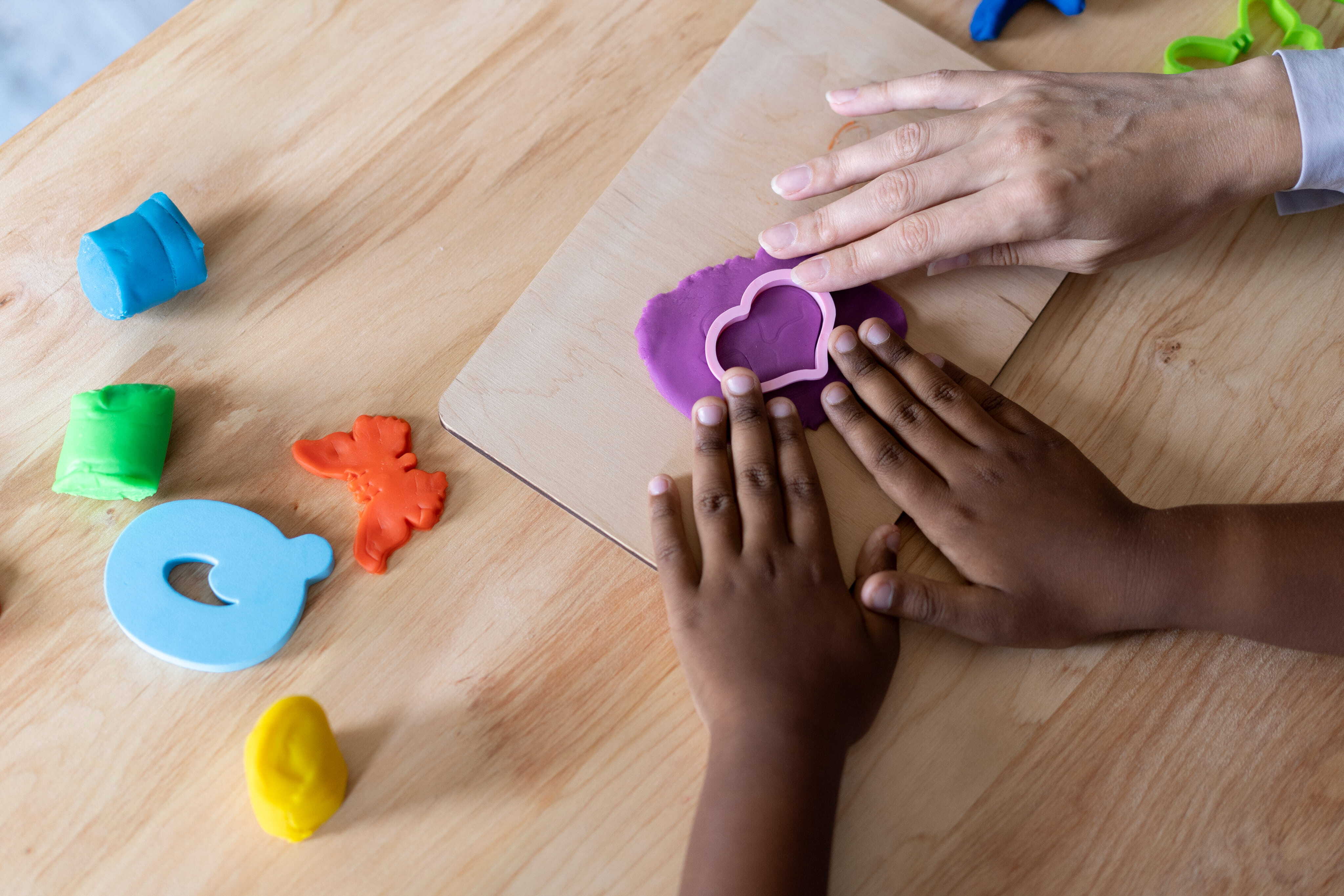 A high-angle, close-up shot of an adult’s and a child’s hands working together on a wooden table. They are using a heart-shaped cutter on purple modeling clay, surrounded by other colorful pieces of playdough.