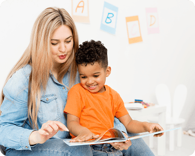 A woman and a young boy sit together, smiling as they read a colorful picture book. In the background, simple "A, B, C, D" posters hang on a white wall, creating a warm learning environment.