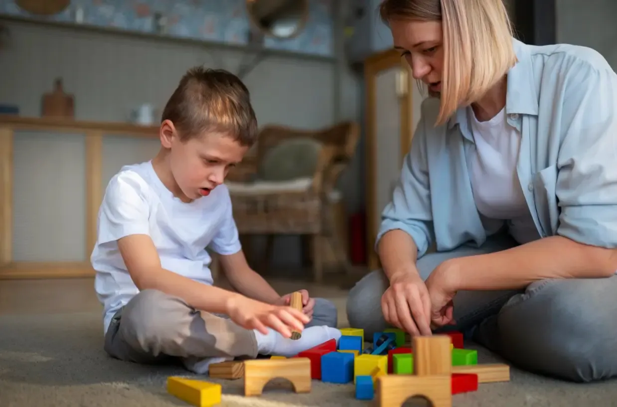 A woman and a young boy sit on the floor, playing with various colorful and natural wooden building blocks on a rug.