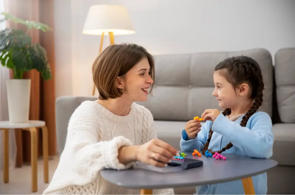 A woman and a young girl with braids smile at each other while playing with colorful plastic connectors at a small table in a living room.