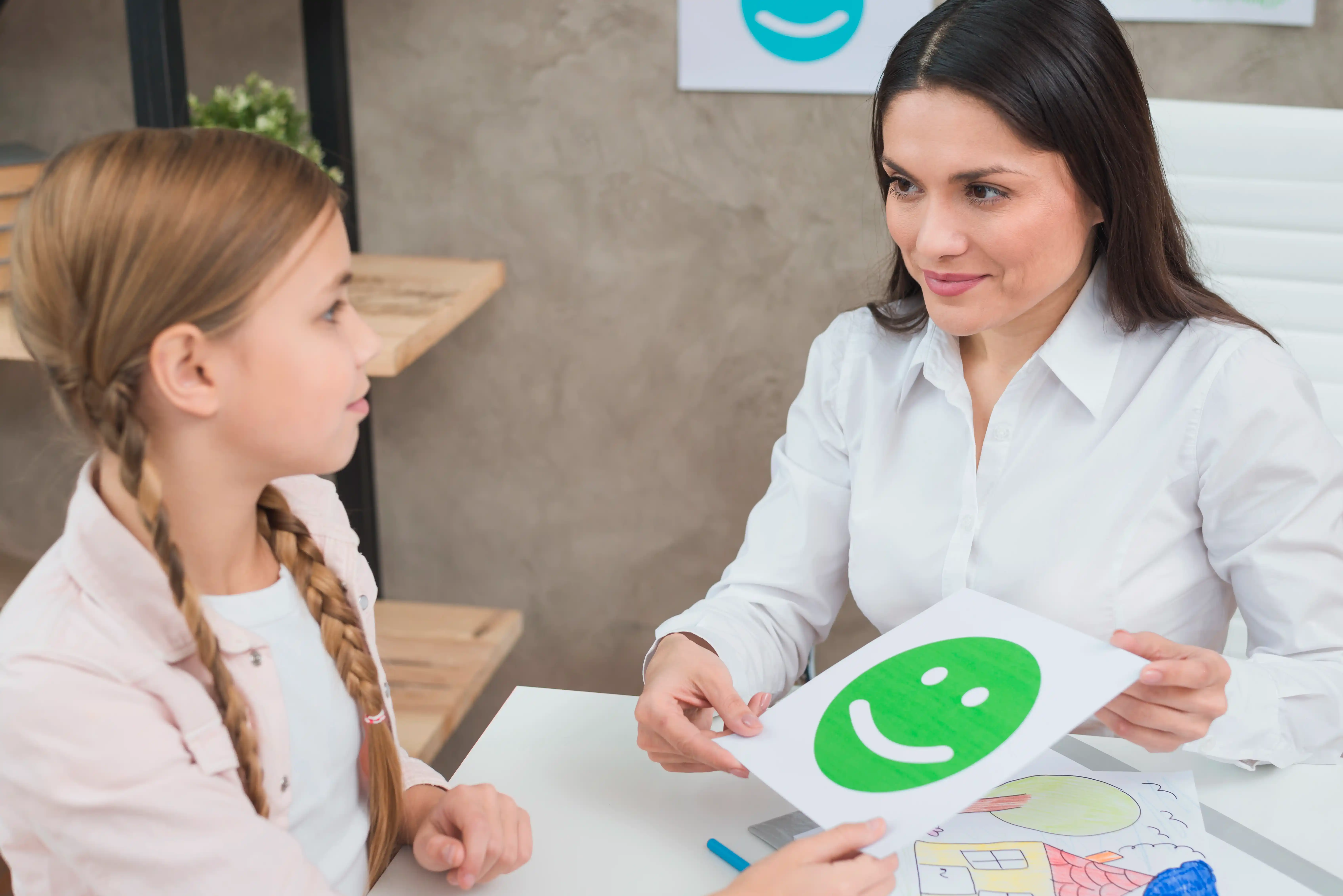A clinician showing a smiley face card to a young girl during a supervised therapy session.