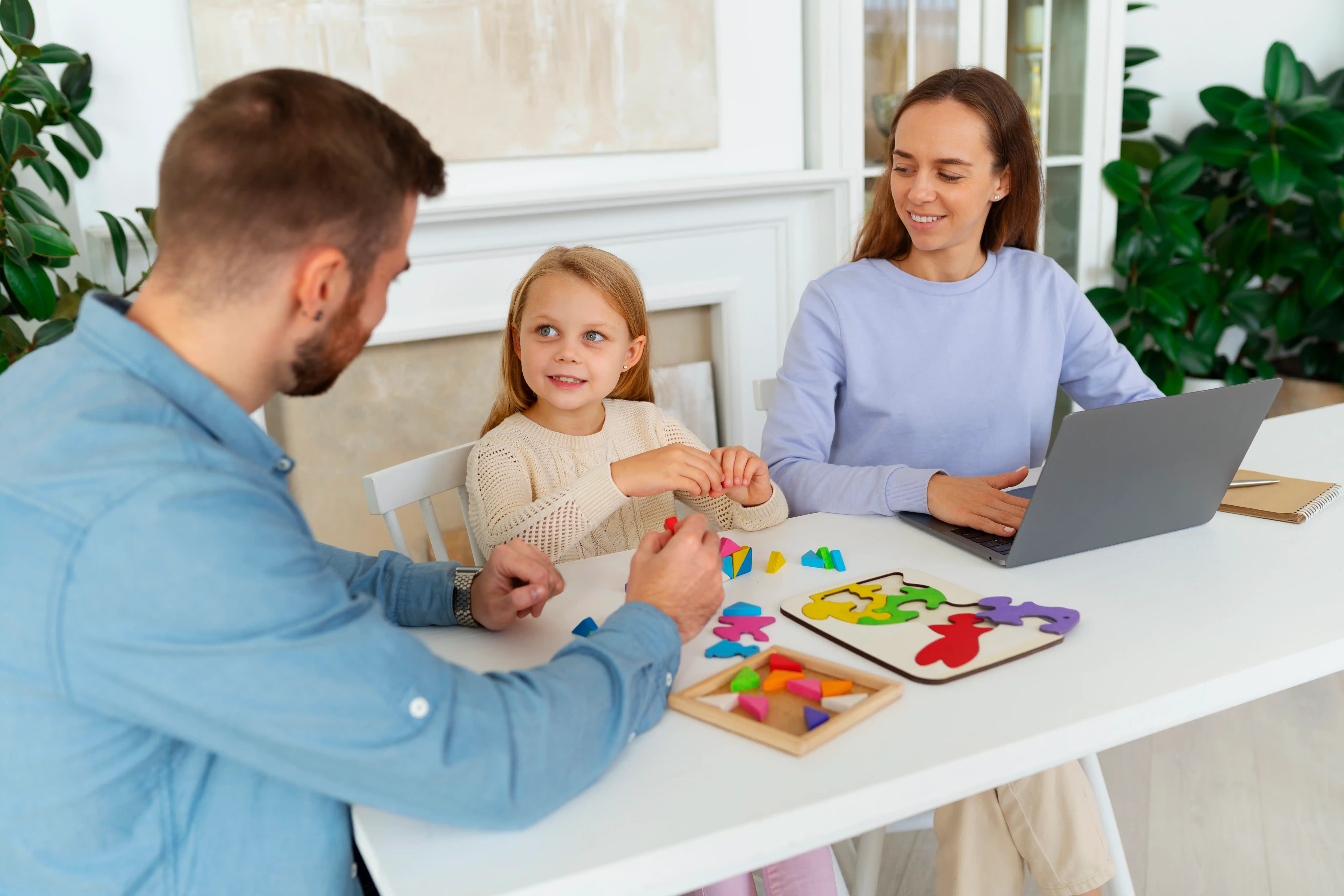 A father and mother sitting at a table with their young daughter, working together on a colorful puzzle activity.